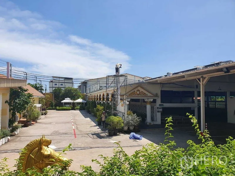 Warehouse for rent in Phra Pradaeng, Samut Prakan - Wide driveway between compound buildings with scaffolding and lush greenery.