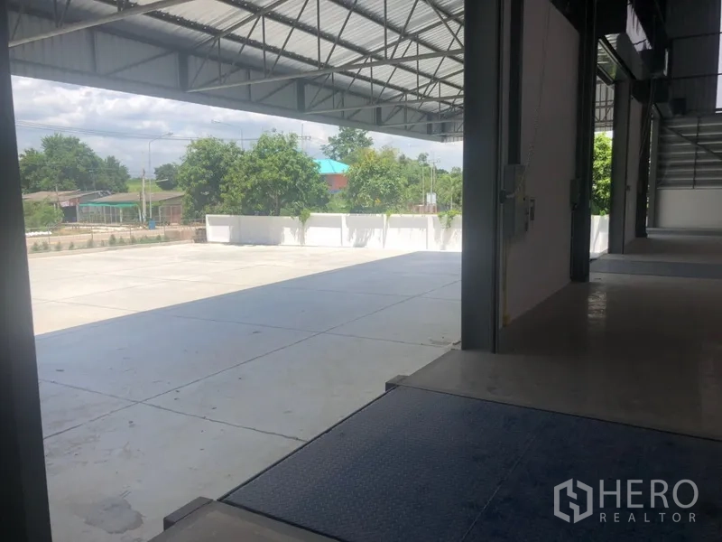 货栈 出租在Si Racha, Chon Buri - 带顶装卸区与坡道，通往大型卡车庭院，围墙外绿树成荫。