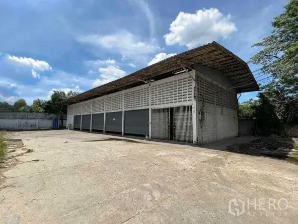 货栈 出租在Bang Na, Bangkok - 曼谷邦纳区单层仓库，多个卷帘门与宽阔水泥前场，蓝天下外观。