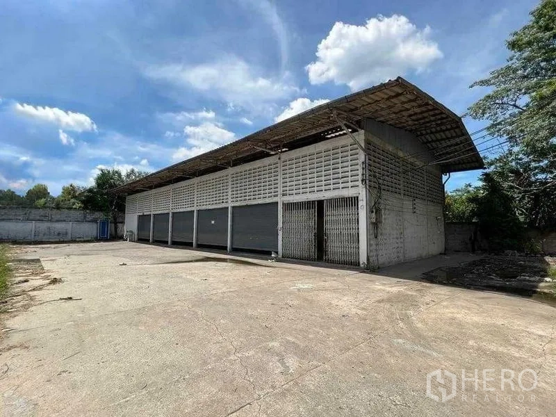 货栈 出租在Bang Na, Bangkok - 曼谷邦纳区单层仓库，多个卷帘门与宽阔水泥前场，蓝天下外观。