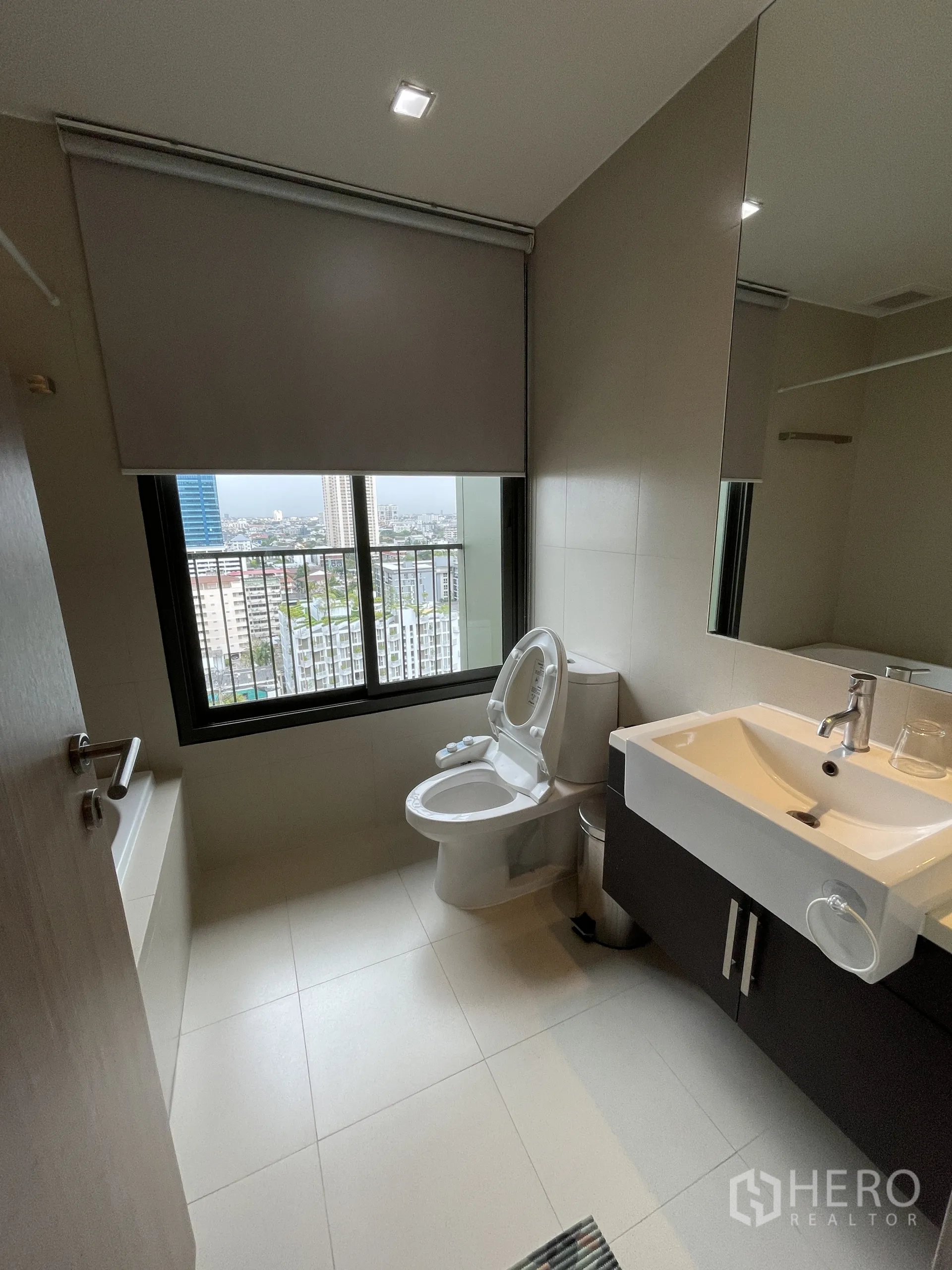 公寓 出租在Watthana, Bangkok - 浴室设大镜面、台盆、坐便器及带城市景观的窗户。