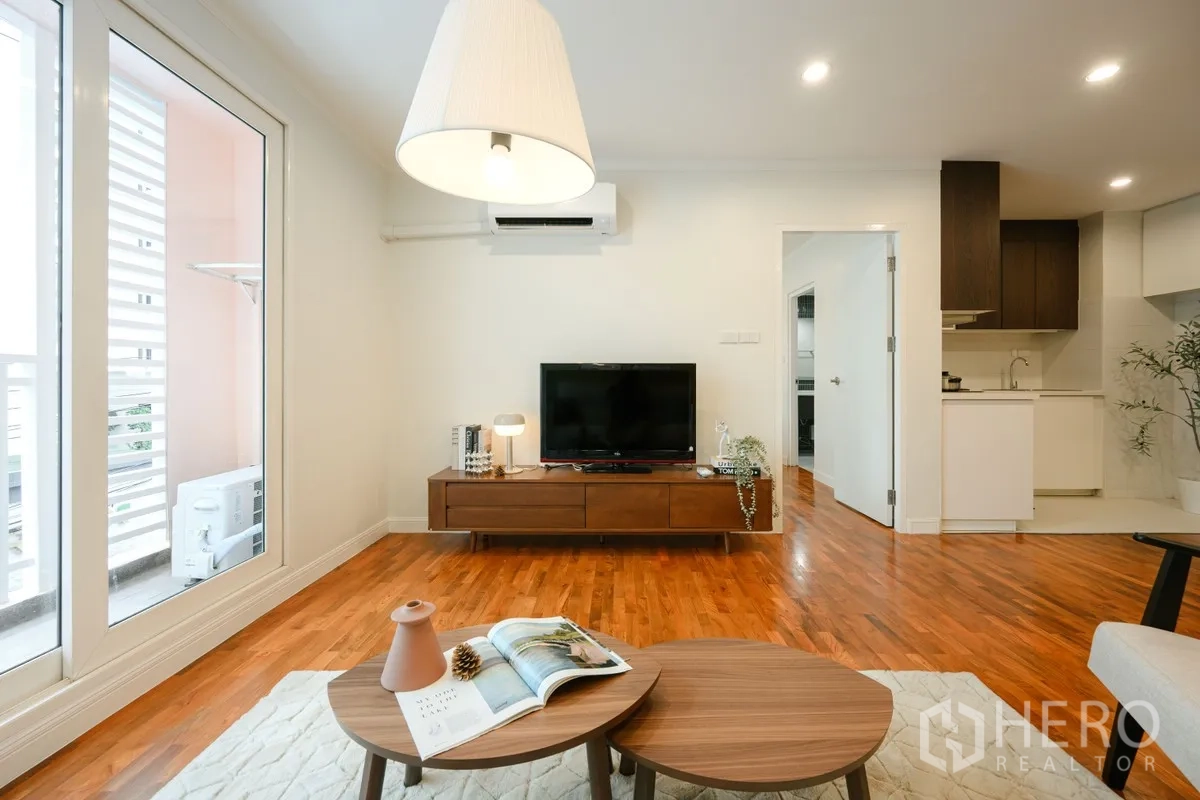 Condominium for sale in Watthana, Bangkok - Living room facing a wooden TV unit, balcony doors, and an adjacent kitchenette.