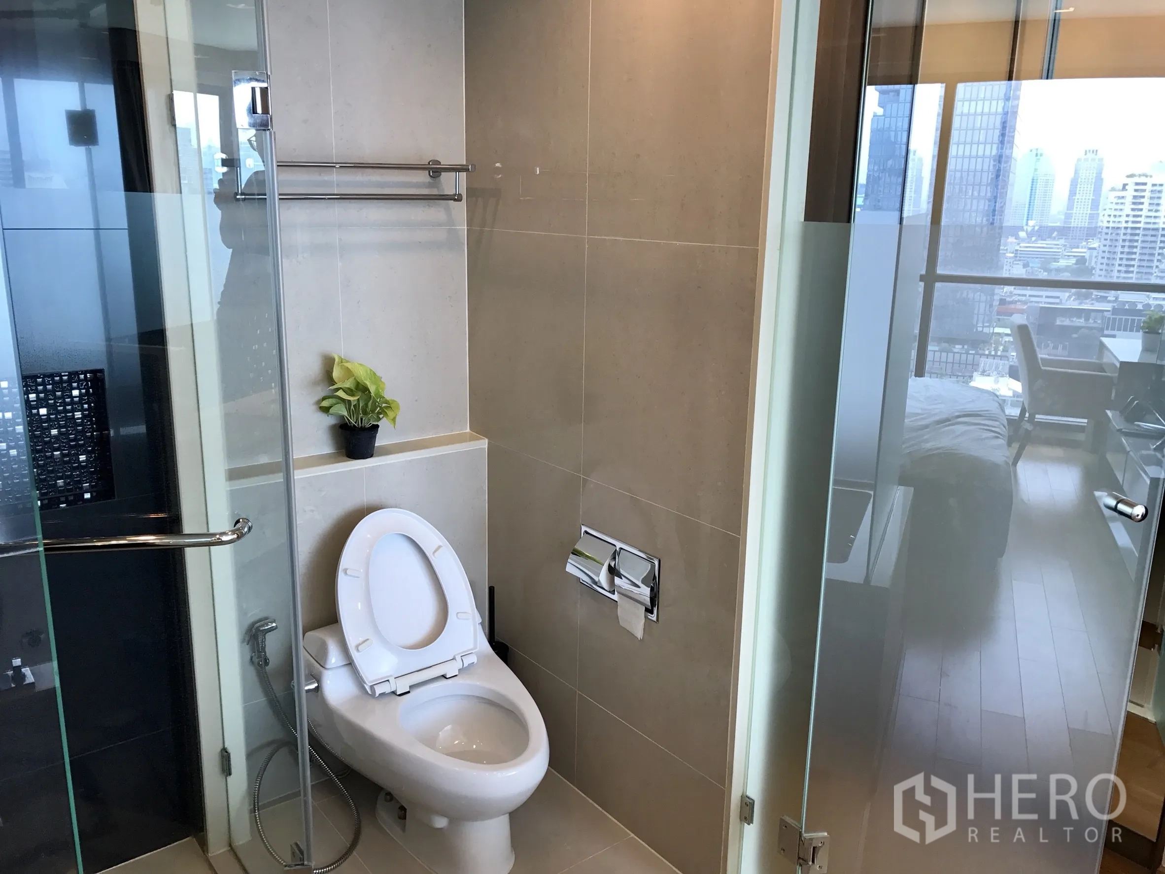 公寓 出租或出售在Bang Rak, Bangkok - 明亮卫生间，玻璃淋浴间与坐便器，门外可见城市景色。