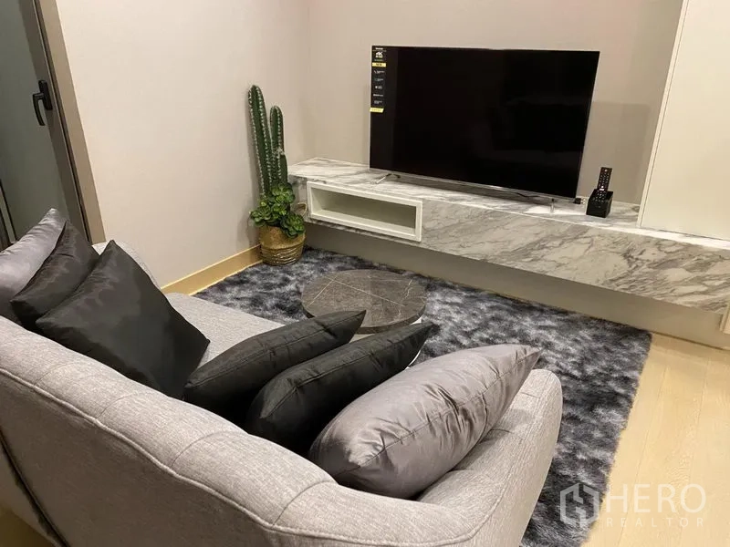 Condominium for rent in Khlong Toei, Bangkok - Modern living room with gray sofa, marble floating TV console, shaggy rug, cactus and round coffee table.