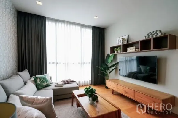 Condominium for rent in Watthana, Bangkok - Cozy living room with L-shaped sofa, large wall-mounted TV, floating shelves and floor-to-ceiling curtained window.