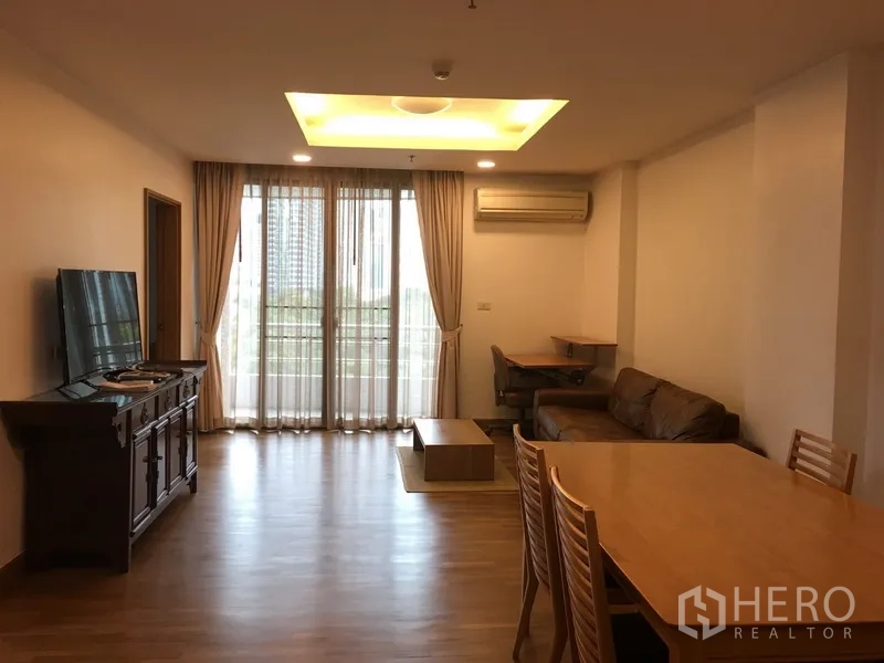 Condominium for rent in Pathum Wan, Bangkok - Bright living room with wood floors, recessed ceiling, TV console, sofa, dining set and full-height balcony doors with city outlook.