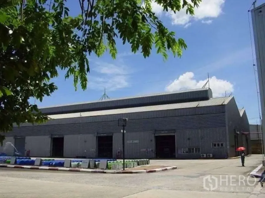 工厂 出售在Rayong - 金属外墙的大型厂房，双坡高屋顶，多个卷帘装卸门与宽阔混凝土场地，前景有树影，蓝天白云。