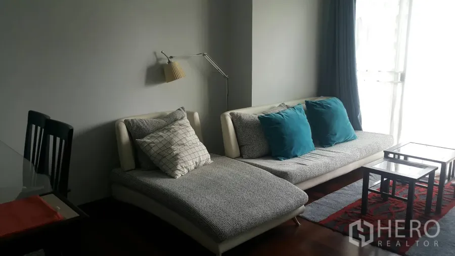 Condominium for rent in Watthana, Bangkok - Bright living room with grey sectional sofa, teal cushions, glass coffee tables and a floor lamp by full-height curtains.