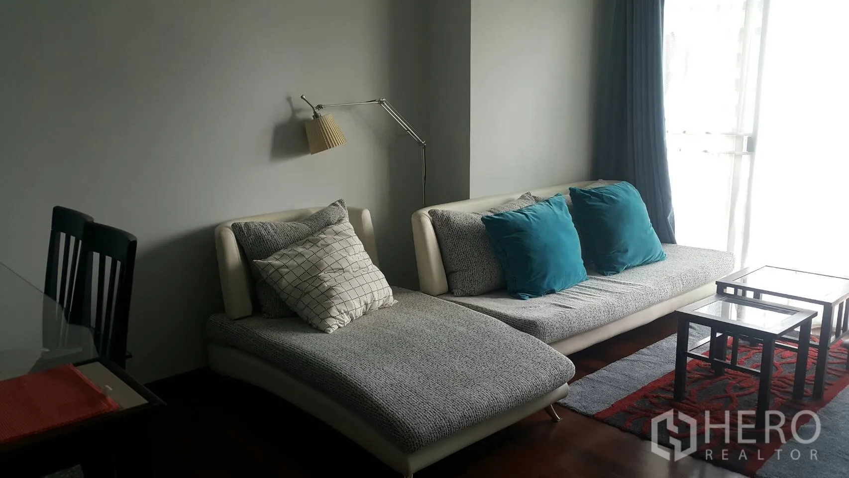 Condominium for rent in Watthana, Bangkok - Bright living room with grey sectional sofa, teal cushions, glass coffee tables and a floor lamp by full-height curtains.