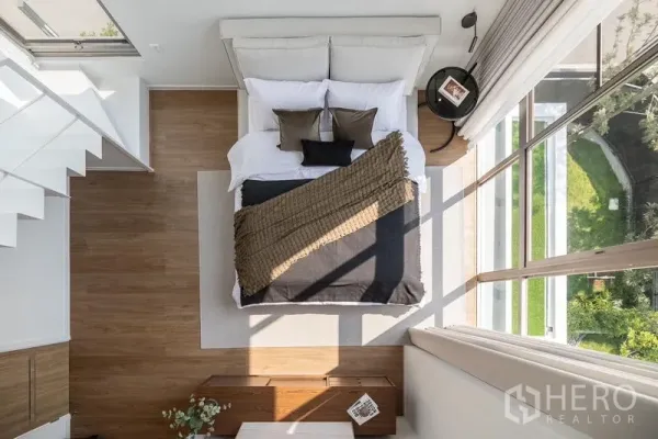 Detached House for rent in Saphan Sung, Bangkok - Top-down view of a cozy bed beside a double-height window with green views and wood floors.