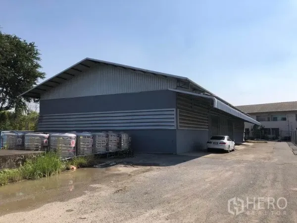 工厂 出租在Bang Bua Thong, Nonthaburi - 灰色金属外立面的工厂，带有遮雨装卸区和停车位，晴天拍摄。