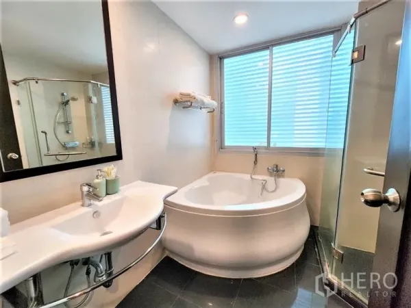 公寓 出租在Bang Rak, Bangkok - 明亮浴室，配圆形浸泡浴缸、玻璃淋浴间、挂墙式洗手台与磨砂窗。