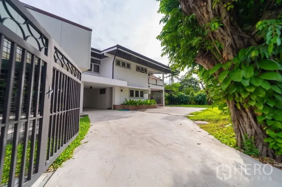 Detached House for rent in Khlong Toei, Bangkok - View from the front gate to the two‑storey home with a long driveway and mature tree.