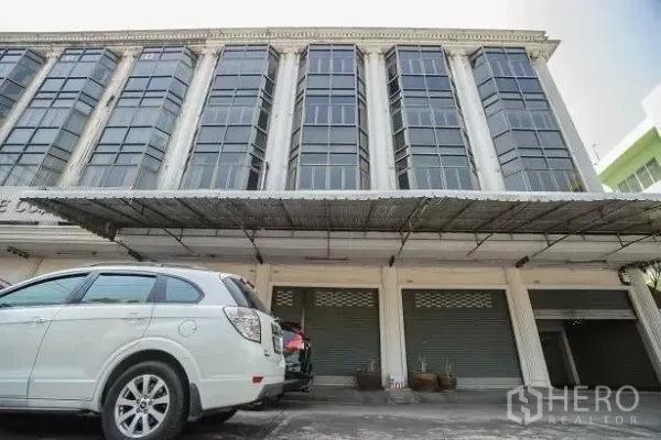 店屋 出租在Wang Thonglang, Bangkok - 建筑立面近景，大面积玻璃、金属雨棚、卷闸门与场内停车位。