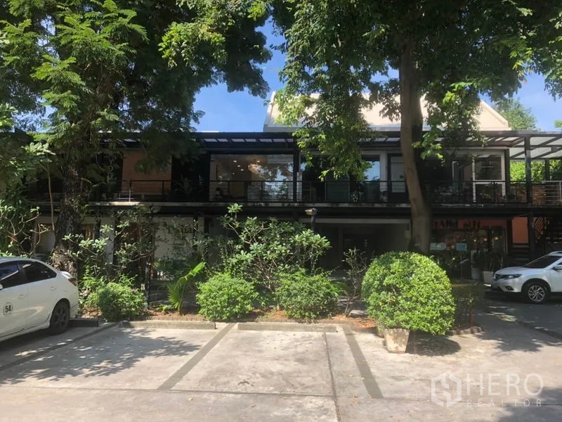 零售空间 出租在Watthana, Bangkok - 低层商业排铺前的遮阴停车位与成排绿树。