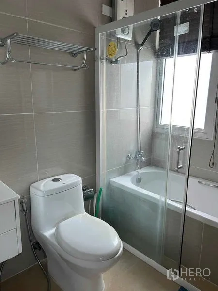 Condominium for sale in Huai Khwang, Bangkok - Modern bathroom with toilet, glass shower over bathtub and window.