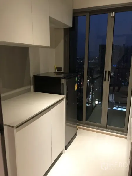 公寓 出租在Phra Khanong, Bangkok - 紧凑厨房，白色橱柜与冰箱，推拉玻璃门通向阳台赏夜景。