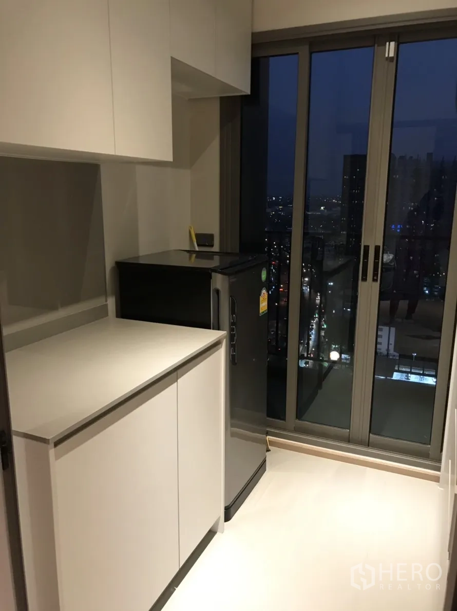 公寓 出租在Phra Khanong, Bangkok - 紧凑厨房，白色橱柜与冰箱，推拉玻璃门通向阳台赏夜景。