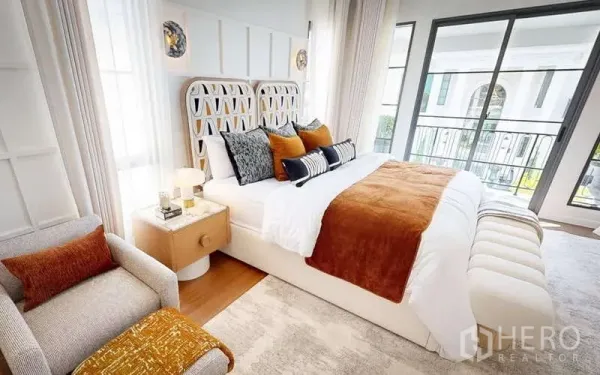 Detached House for rent in Bang Phli, Samut Prakan - Sunlit bedroom with balcony access, patterned headboard, warm orange accents and a bench at the foot of the bed.