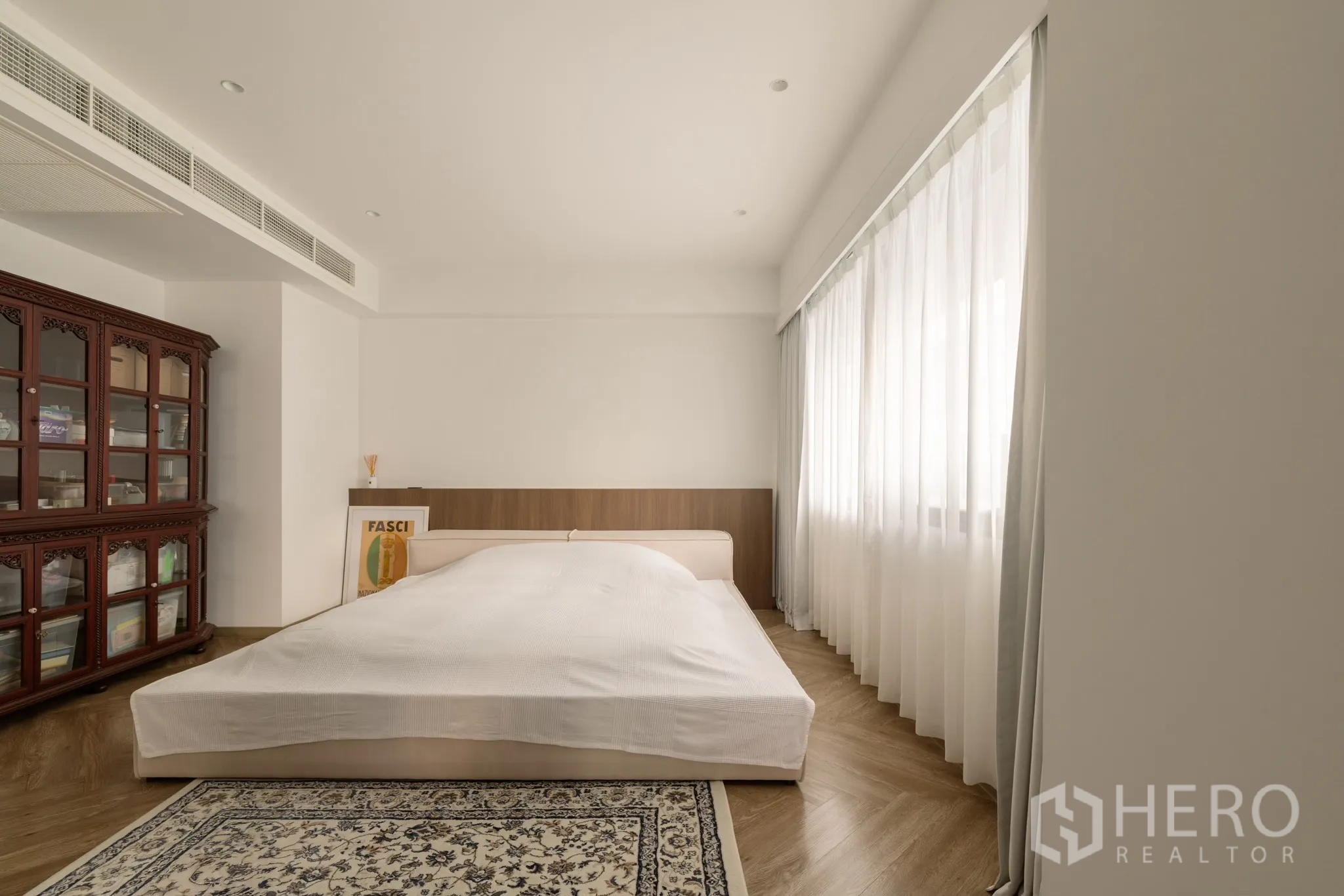 Condominium for sale in Khlong Toei, Bangkok - Minimal master bedroom with low platform bed, large window with sheer curtains and ornate wooden cabinet.