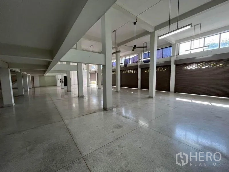 店屋 出租在Khlong Toei, Bangkok - 一层大厅层高高，带吊扇与夹层，多组金属卷帘门便于装卸。