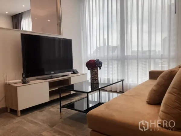 公寓 出租在Sathon, Bangkok - 明亮客厅，电视置于矮柜上，玻璃茶几，落地纱帘可见城市景观。