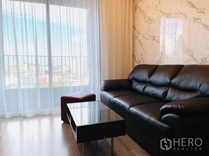 公寓 出租在Bang Na, Bangkok - 客厅配黑色皮沙发、茶几与大理石质感背景墙，纱帘外为城市景观。