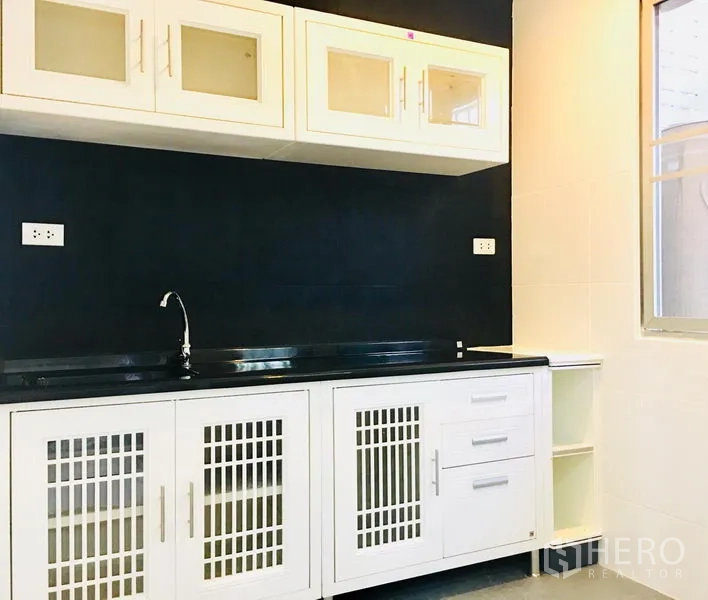 Townhouse for rent in Prawet, Bangkok - Fitted kitchen with white cabinetry, black countertop, sink, and a window against dark tiled walls.