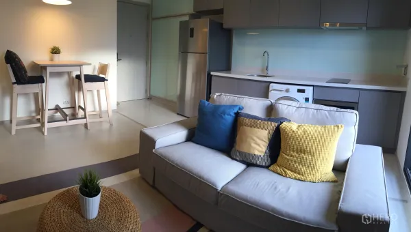 公寓 出租在Khlong Toei, Bangkok - 开放式客厅，灰色沙发与彩色抱枕，吧台餐桌，配备冰箱和洗衣机的现代厨房。