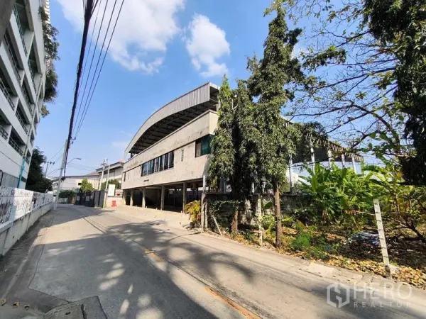 Warehouse for rent in Huai Khwang, Bangkok - Street view of a concrete industrial building with curved roof and greenery in Huai Khwang, Bangkok.