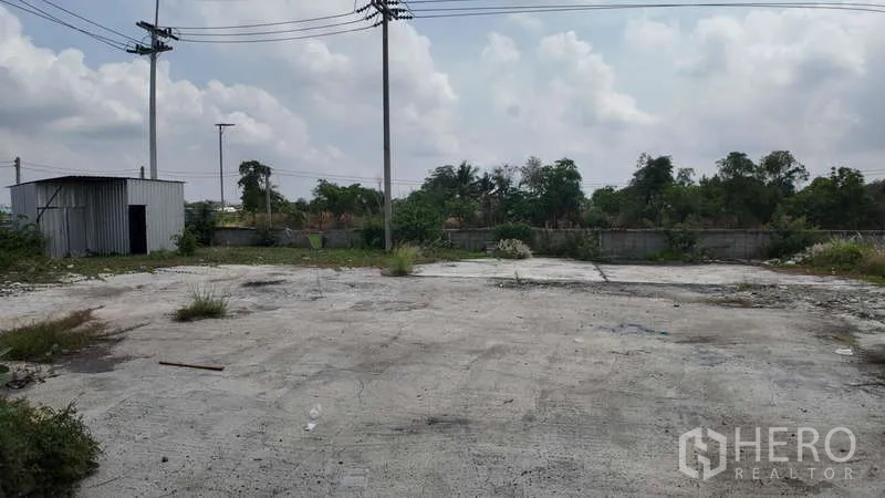 Warehouse for rent in Chachoengsao - Open yard with rough concrete surface, utility poles and a small shed at the boundary.