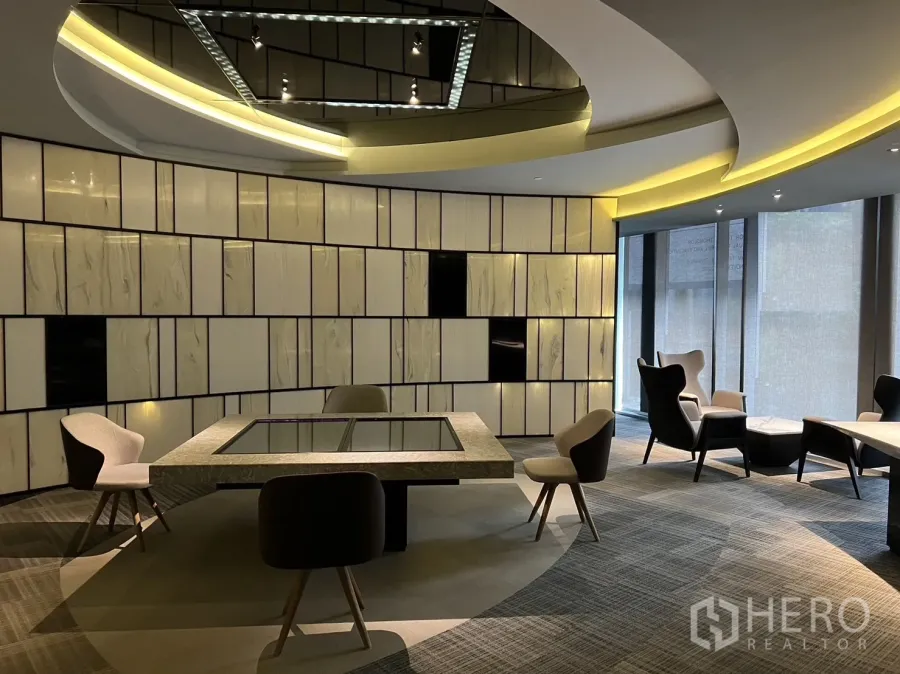 零售空间 出租在Watthana, Bangkok - 现代零售展示空间，弧形吊顶灯带、几何装饰墙、地毯地面与中央展示桌及休闲座椅。