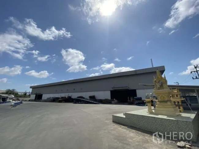 Warehouse for rent in Mueang Samut Sakhon, Samut Sakhon - Metal-clad warehouse exterior with expansive paved yard and a spirit house at the entrance.