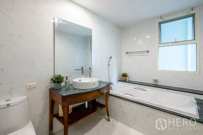 Condominium for sale in Watthana, Bangkok - White marble-style bathroom with bathtub, vanity and window for natural light.