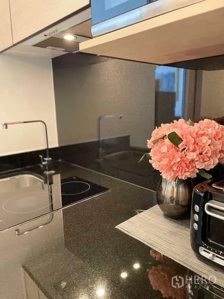 Condominium for rent or sale - Modern kitchenette featuring black stone countertop, induction hob, sink and small appliances with decorative flowers.