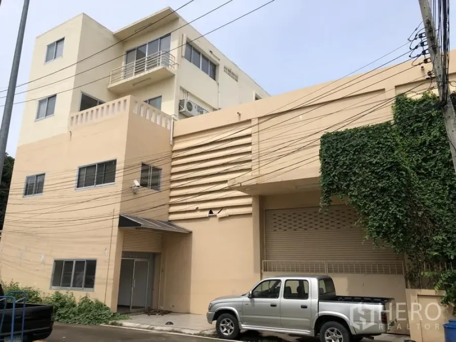 工厂 出租或出售在Bang Lamung, Chon Buri - 米色工业建筑外立面，带装卸区、卷闸门和停靠的皮卡车。