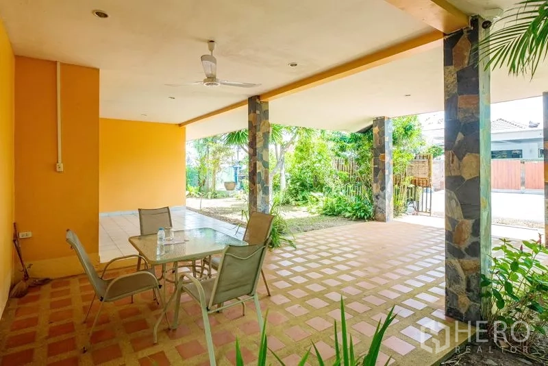 Detached House for sale in Bang Lamung, Chon Buri - Covered outdoor terrace with ceiling fan, dining set, and stone-clad pillars amid greenery.