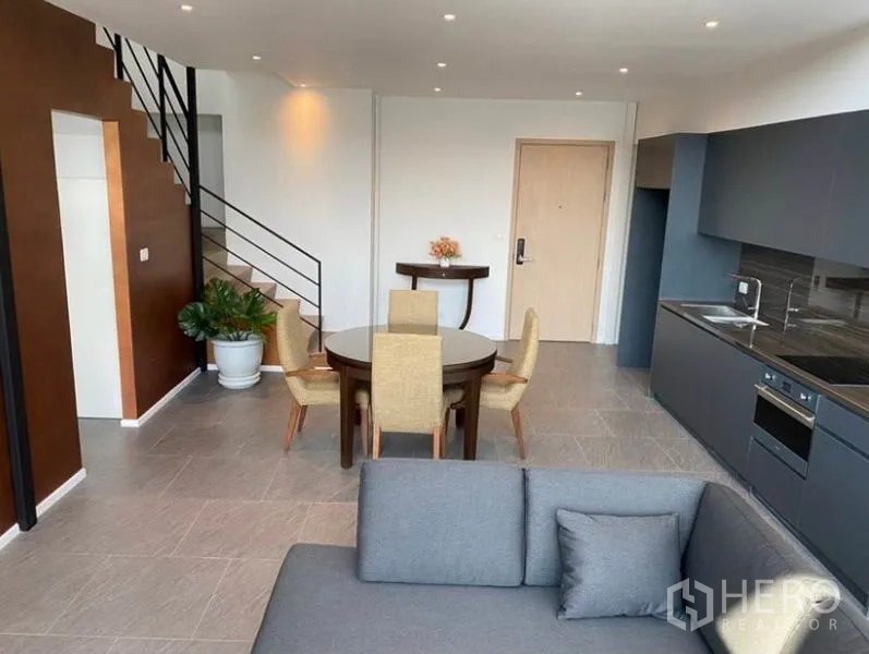 公寓 出租在Bang Rak, Bangkok - 开放式厨房，灰色橱柜、嵌入式烤箱、圆形餐桌与通往夹层的楼梯。