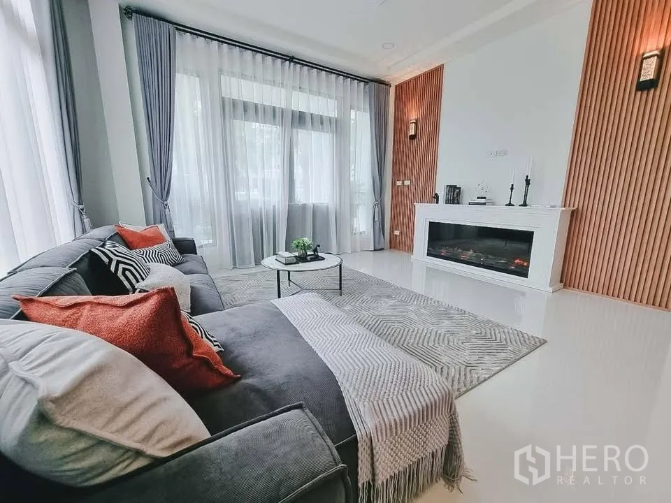 Detached House for rent or sale in Bang Phli, Samut Prakan - Bright living room with large windows, sheer curtains, gray sectional sofa, and a modern fireplace.