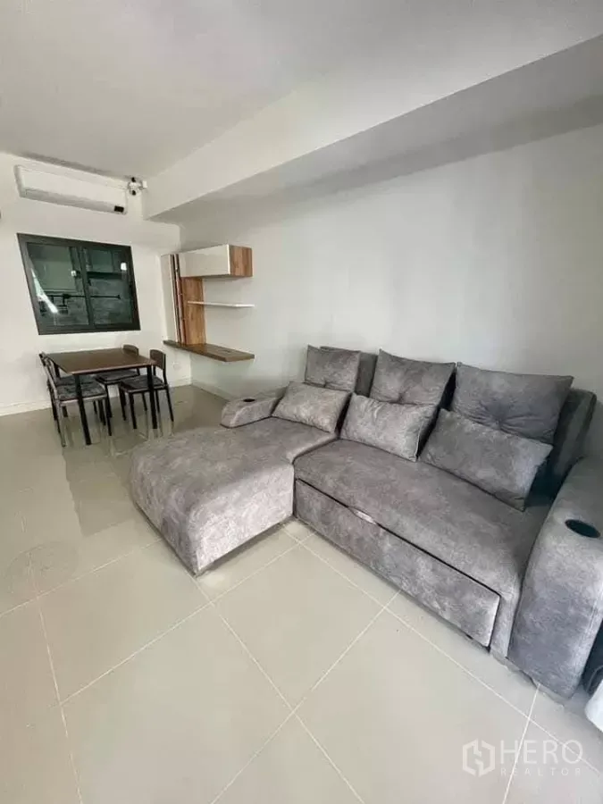 Townhouse for rent in Bang Phli, Samut Prakan - Bright living room with large gray sectional sofa, tiled floor, dining table and floating shelves.