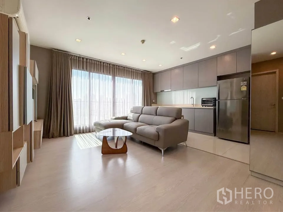 Condominium for rent in Khlong Toei, Bangkok - Open living room with large windows, sofa and one-wall kitchen with fridge.