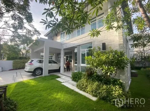 Detached House for rent in Prawet, Bangkok - Side view showing the covered carport and landscaped lawn with mature trees and shrubs.