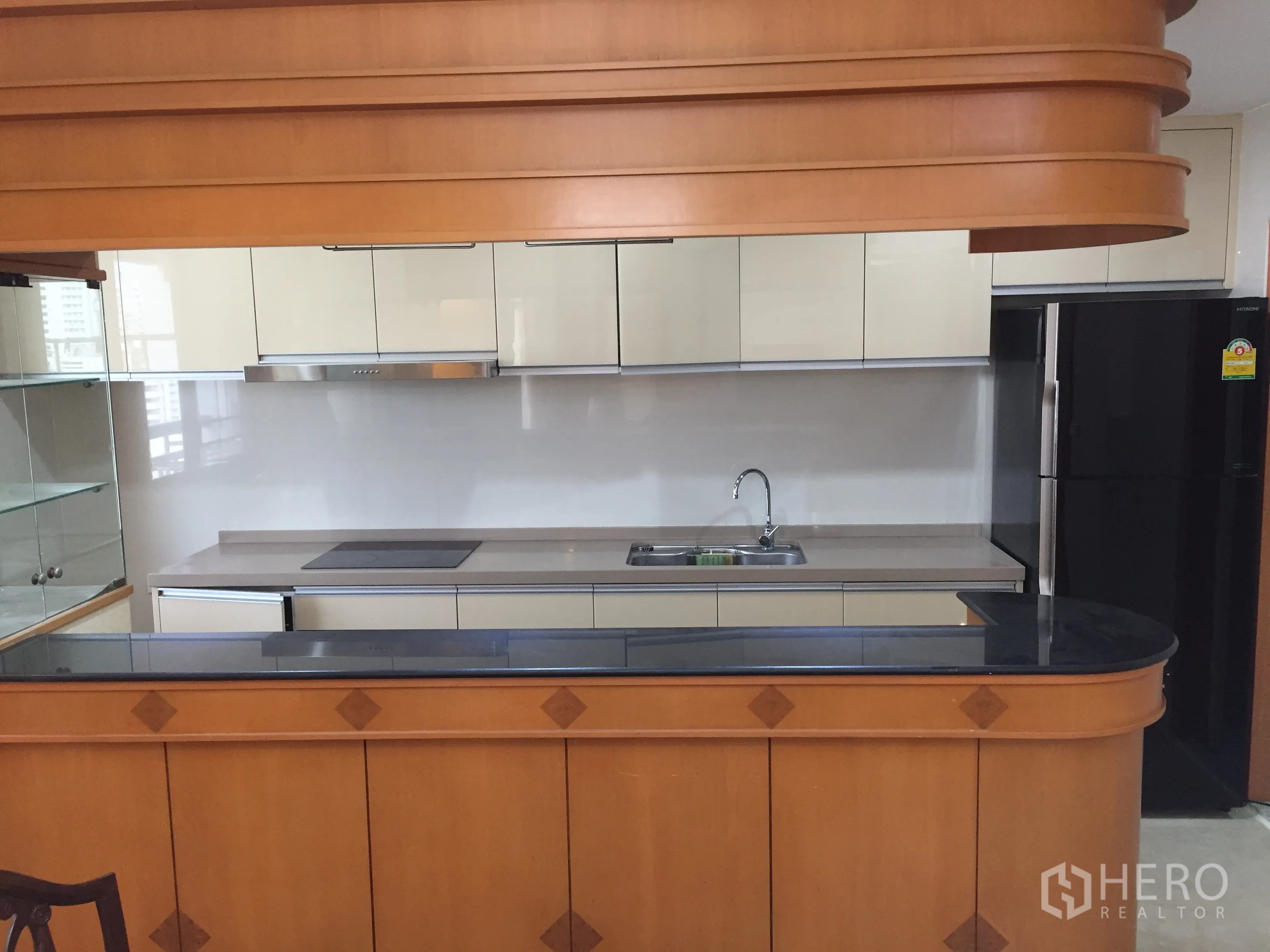Condominium for rent in Watthana, Bangkok - Full-size fitted kitchen with glossy cabinets, electric cooktop, sink, and a curved wood breakfast bar with black countertop.