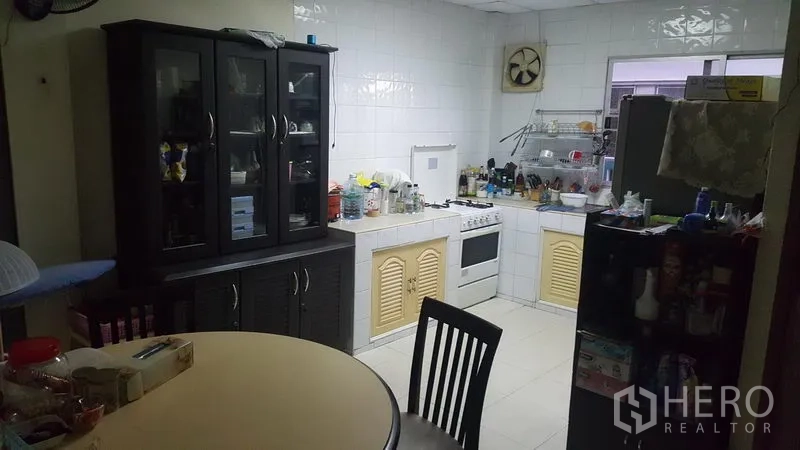 Shophouse for sale in Khlong Toei, Bangkok - Tiled kitchen with gas stove, cabinets, dining table and open shelving beside a window.