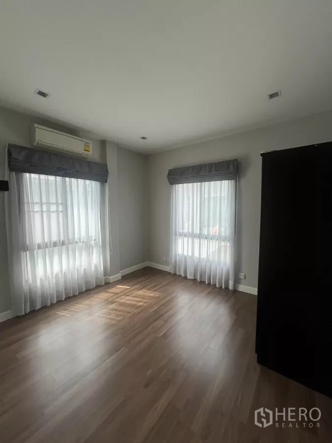 Detached House for sale in Bang Kapi, Bangkok - Sunlit bedroom with two corner windows, air conditioner, and wood floors.