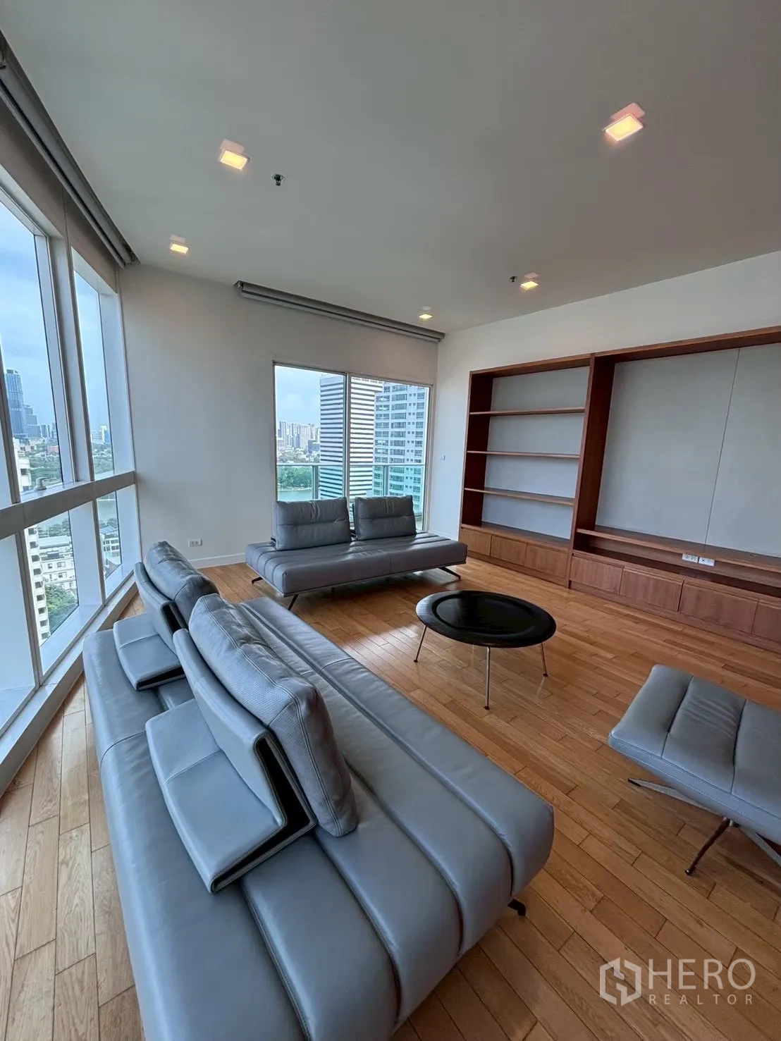 Condominium for rent in Khlong Toei, Bangkok - Corner lounge with panoramic windows, city views, wood floors, and wall shelving.