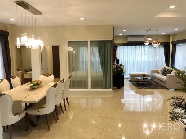 Detached House for rent or sale in Saphan Sung, Bangkok - Dining area with long table and pendant lights beside a bright lounge with floor-to-ceiling curtains.