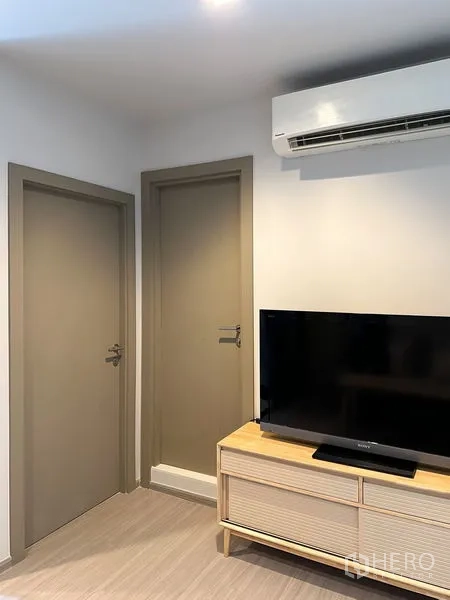 Condominium for sale in Ratchathewi, Bangkok - Compact living area with wall-mounted air conditioner, two interior doors, and a TV on a wooden console.