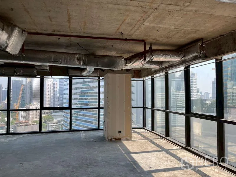 Office Space for rent - Sunlit corner office shell with full-height glazing and concrete ceiling overlooking city buildings.