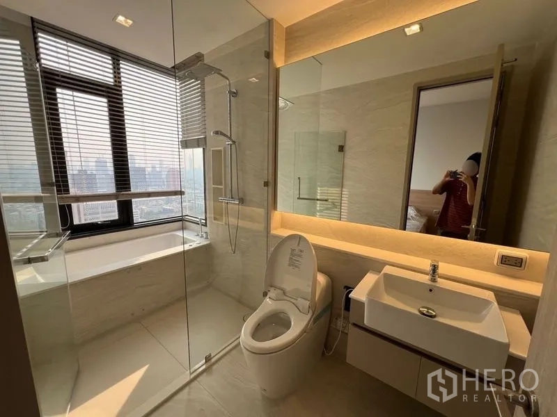 คอนโด ให้เช่าในRatchathewi, Bangkok - ห้องน้ำพร้อมอ่างอาบน้ำริมหน้าต่าง ฉากกั้นอาบน้ำกระจก อ่างล้างหน้า และโถสุขภัณฑ์ มองเห็นวิวเมือง